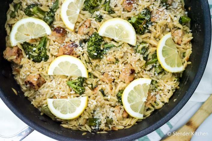 Healthy Lemon Orzo with broccoli, lemon wedges, and chicken sausage in a pan with green napkin.