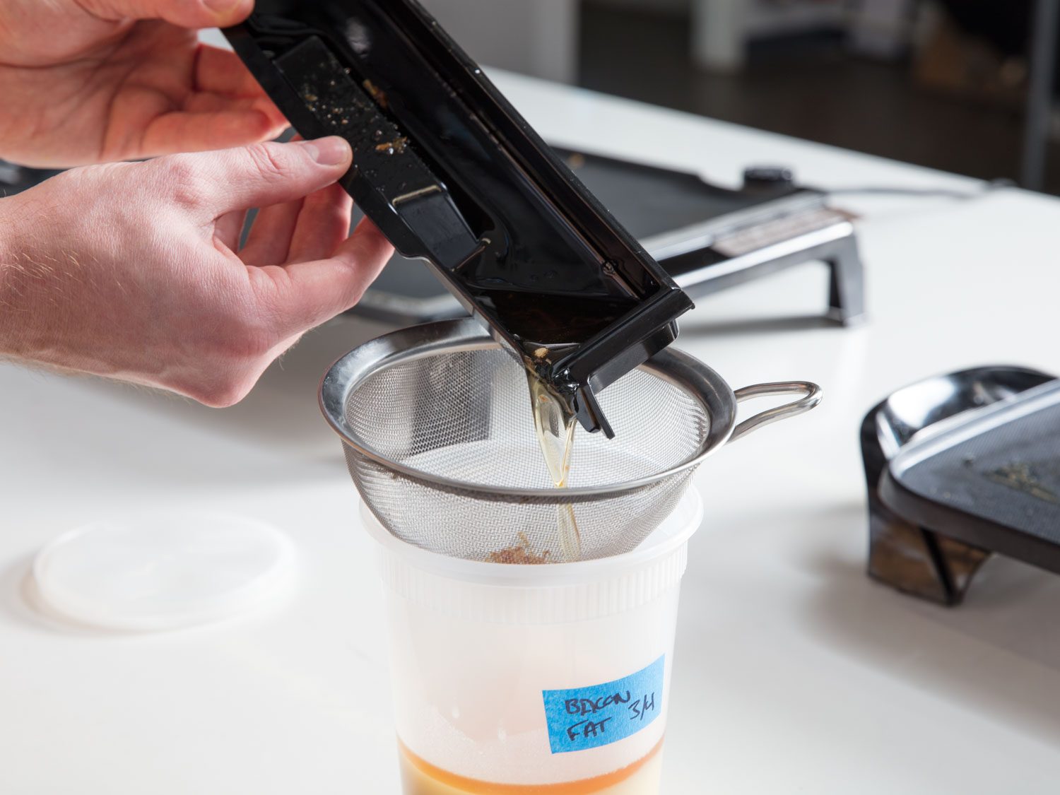 Fat being poured from a grease trap through a fine-mesh strainer into a plastic container labeled 