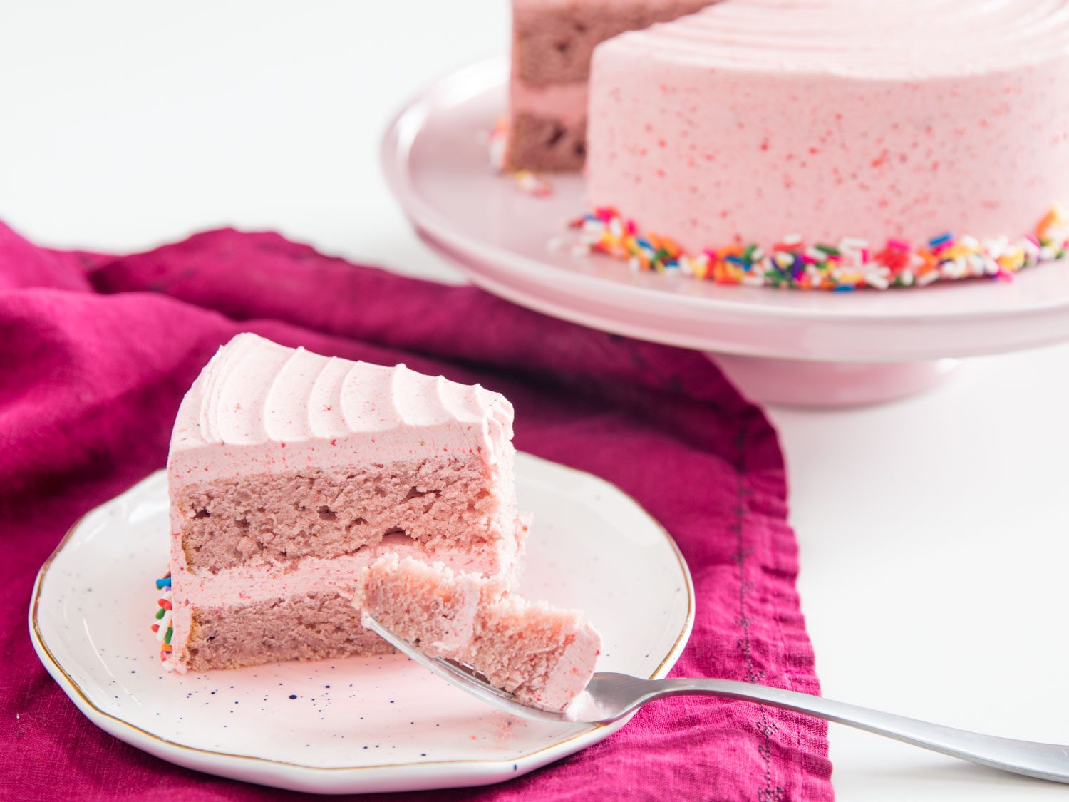 A slice of double-strawberry cake with strawberry frosting, next to the rest of the cake on a cake stand