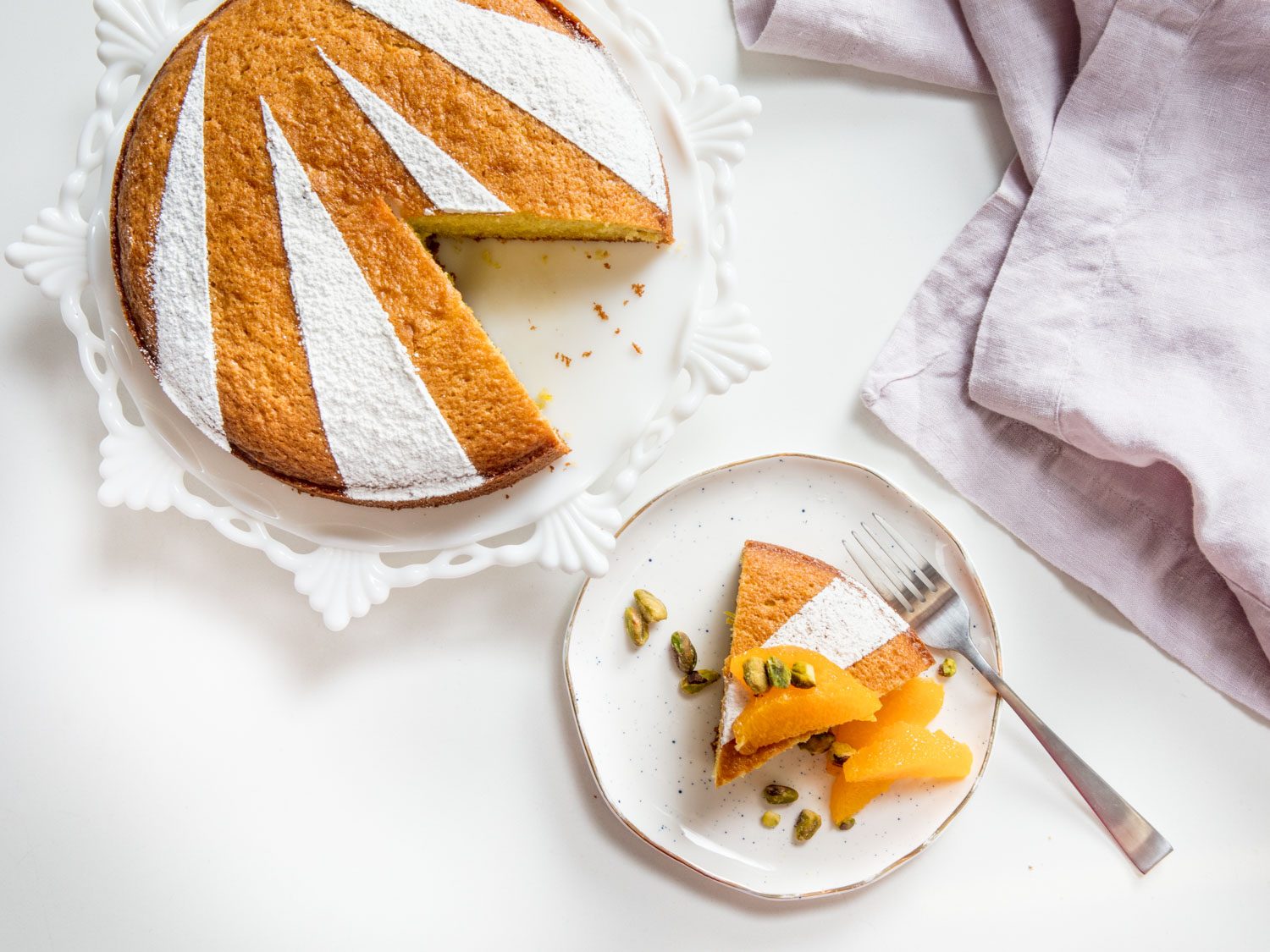 Overhead shot of a slice of olive oil cake on a plate, topped with fruit and nuts, next to the rest of the cake on a cake stand