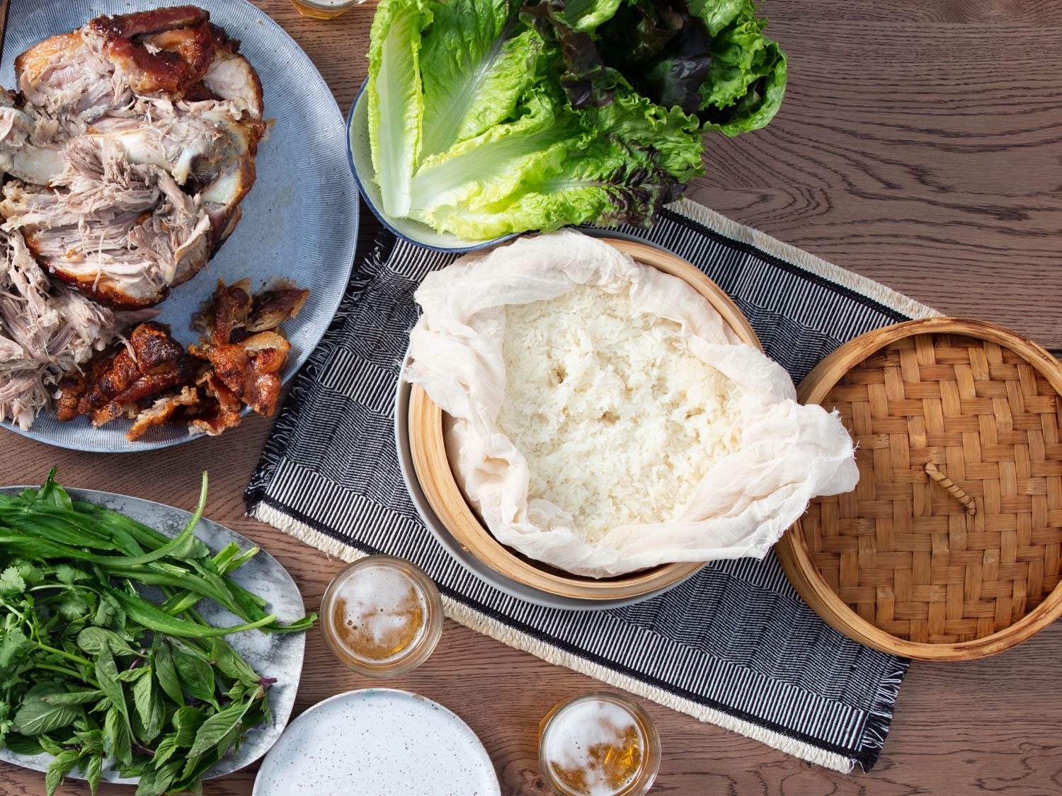 Overhead shot of a steamer basket full of sticky rice alongside a spread of roast pork shoulder, Thai herbs, and lettuces.