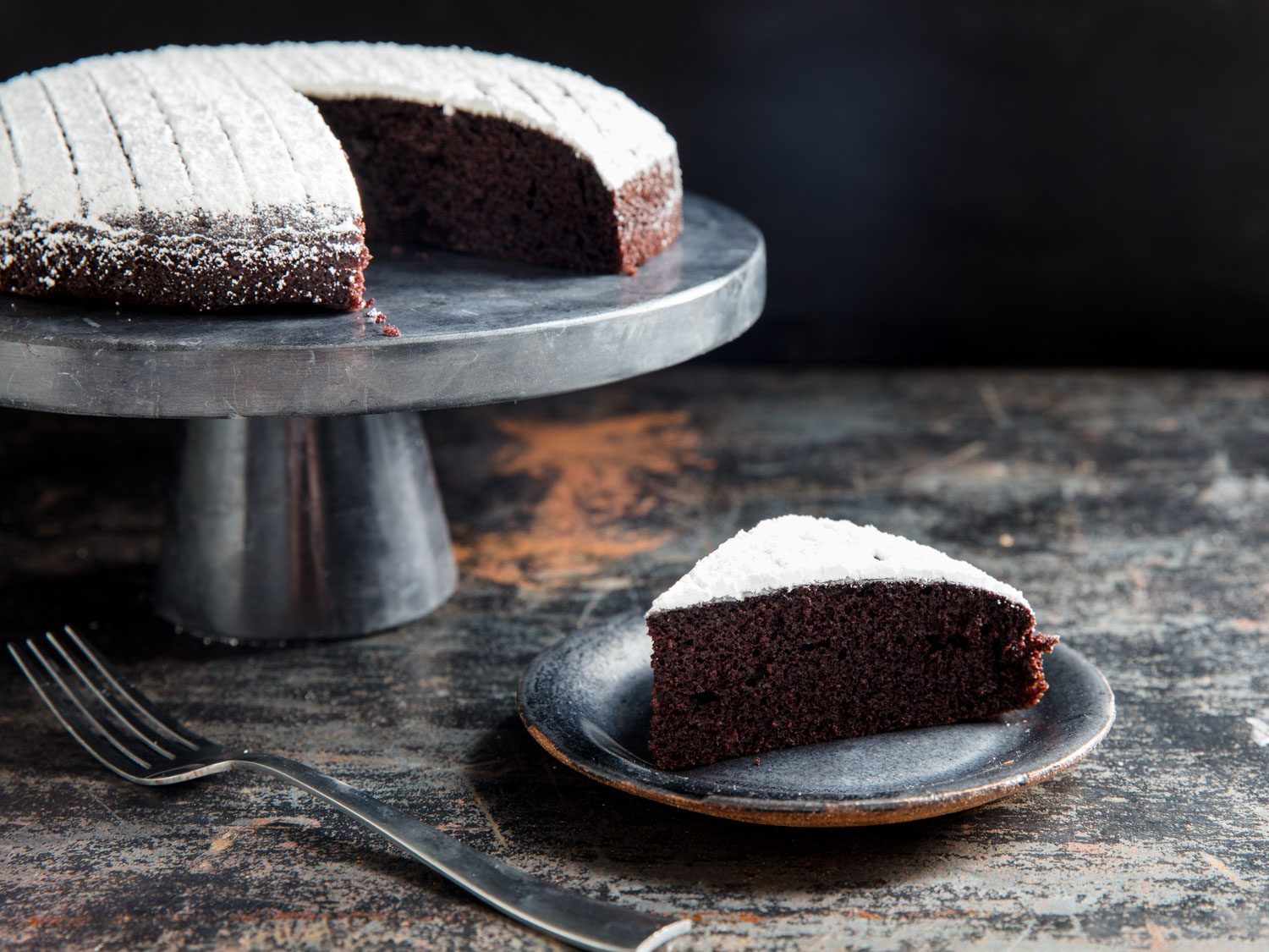 A slice of chocolate olive oil cake on a plate, next to the full cake on a cake stand