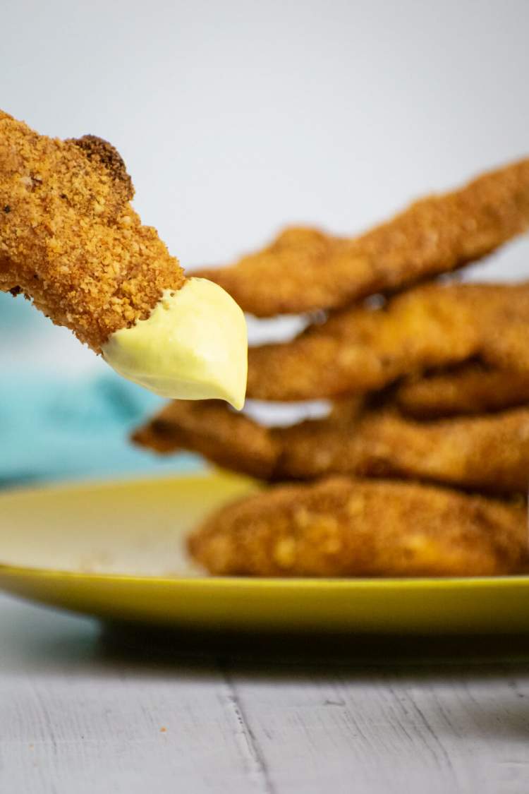 Almond crusted chicken tenders being dipped in honey mustard with chicken tenders on a plate.