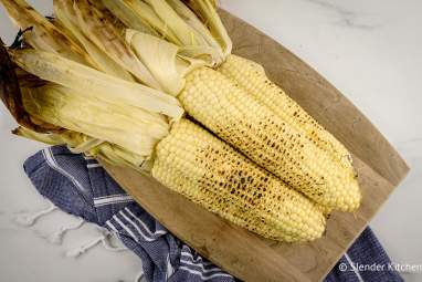 Easy Grilled Corn (How to Grill Corn) Grilled corn with the husks attached piled on a wood cutting board.