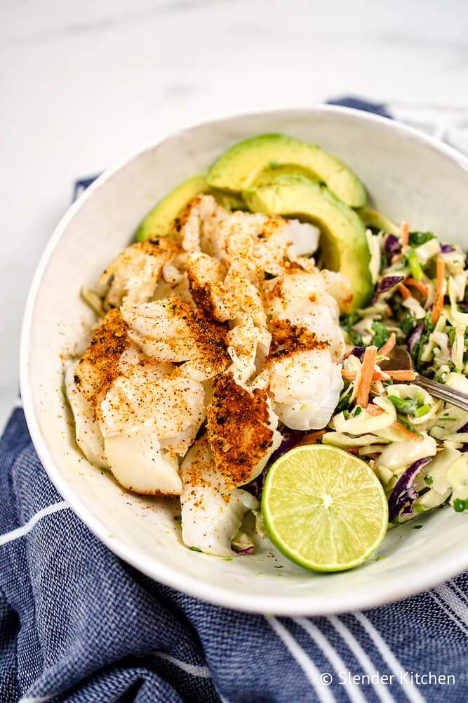 Ancho fish taco bowls with white fish, coleslaw, limes, and avocado in a white bowl.