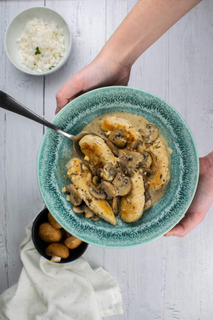 Creamy mushroom chicken in a bowl being held up by two hands alongside some rice and fresh mushrooms.