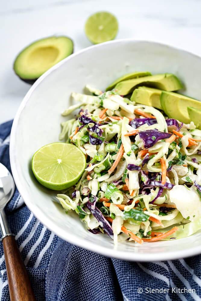 Mexican slaw with avocado, cilantro, cabbage, carrots, and green onions in a white bowl with a wooden spoon.