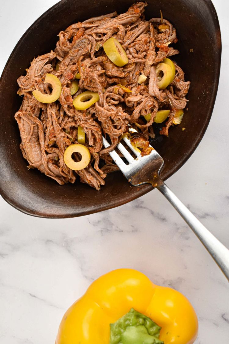 Crockpot ropa vieja in a black dish with shredded beef, peppers, onions, tomatoes, and olives.