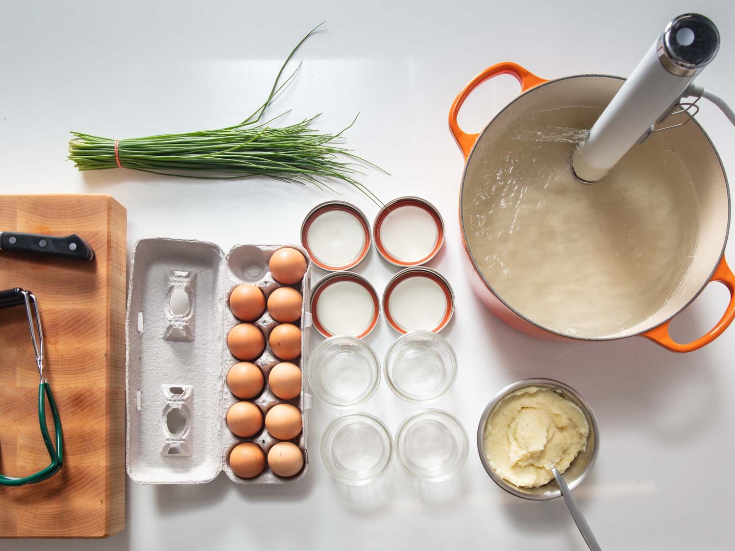 Overhead of set-up and ingredients for sous vide mashed potato egg jars, with an immersion circulator water bath, Mason jars, mashed potatoes, a carton of eggs, and chives.