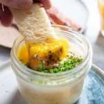 Closeup side view of toast being dipped into a potato-egg jar, breaking the custardy egg yolk.