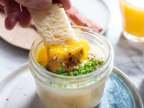Closeup side view of toast being dipped into a potato-egg jar, breaking the custardy egg yolk.