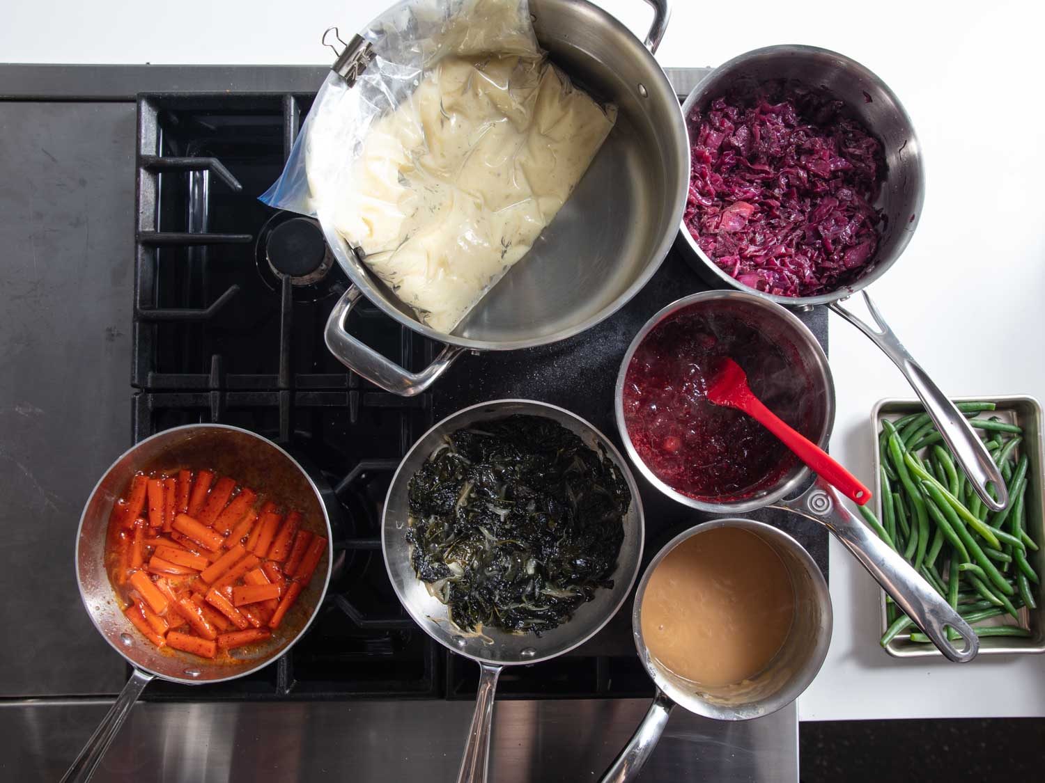 Overhead of a range with Baking Steel set up as a warming station with pots and saucepans of mashed potatoes, gravy, cranberry sauce, braised kale, red cabbage, and glazed carrots.