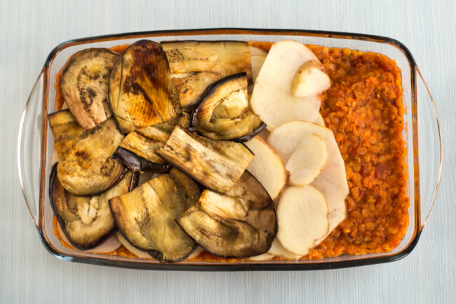 A vegetarian moussaka being layered up in a baking dish with lentils, potatoes and grilled aubergine (eggplant).