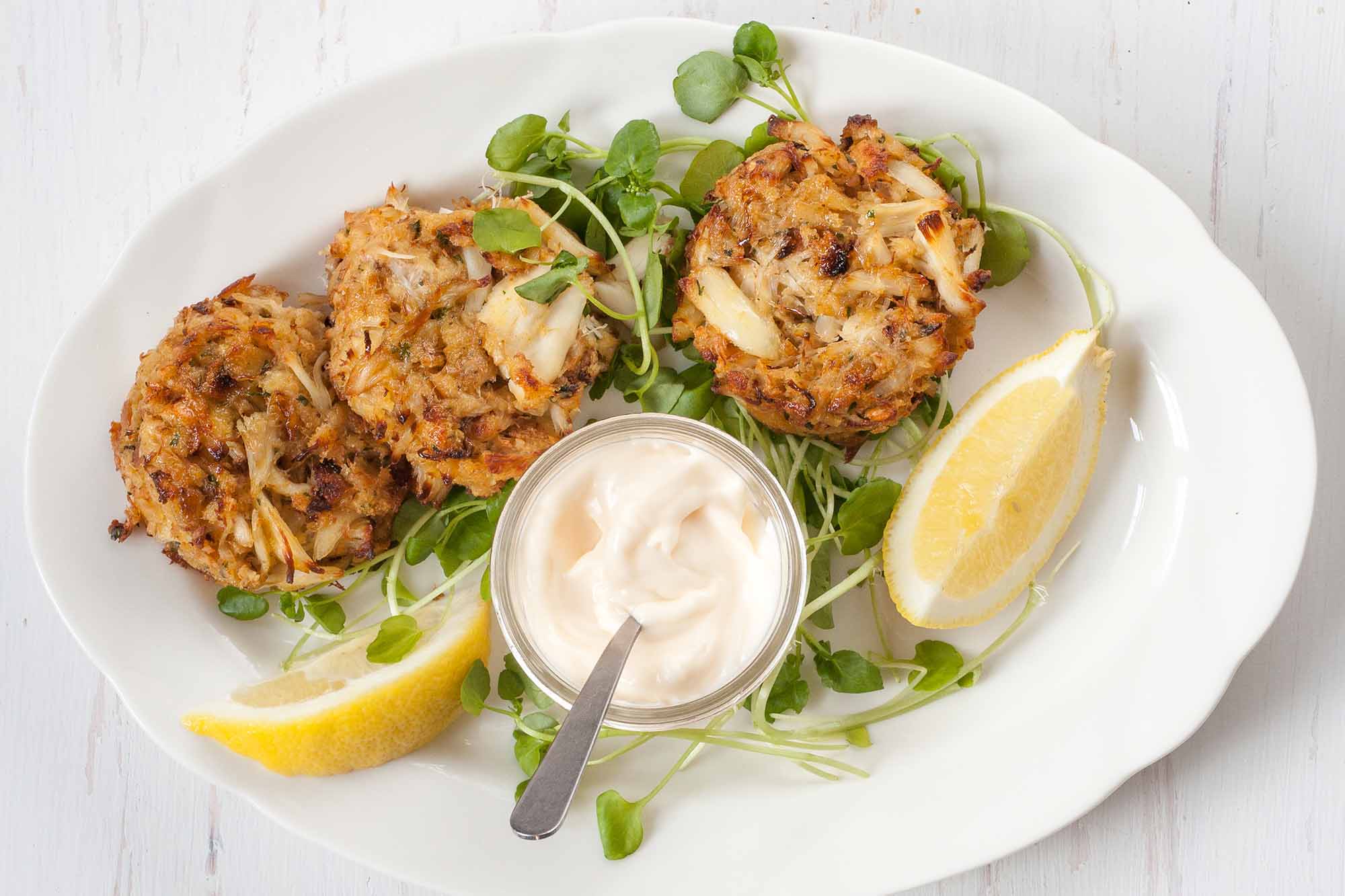 Maryland Crabcakes wtih greens and lemon wedges on a white plate wtih mayo