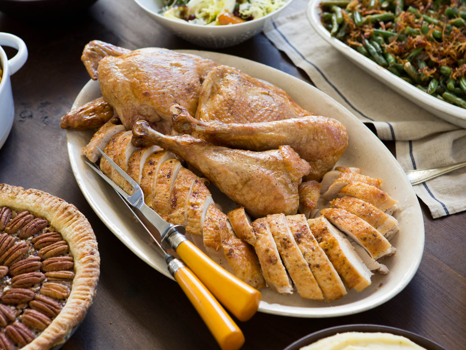 A platter of carved turkey with a carving knife and fork, surrounded by pecan pie, green bean casserole, and salad