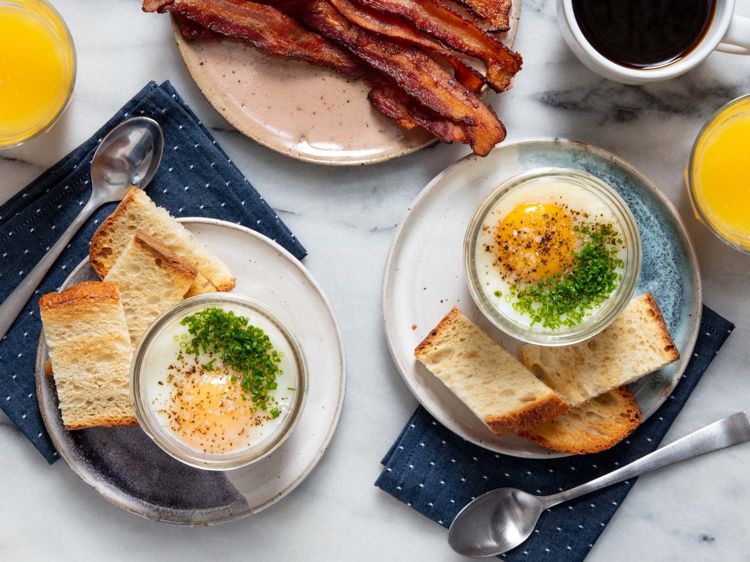 Overhead of two potato-egg jars on plates with toast, and a plate of bacon in the background, along with a glasses of orange juice and a cup of coffee.