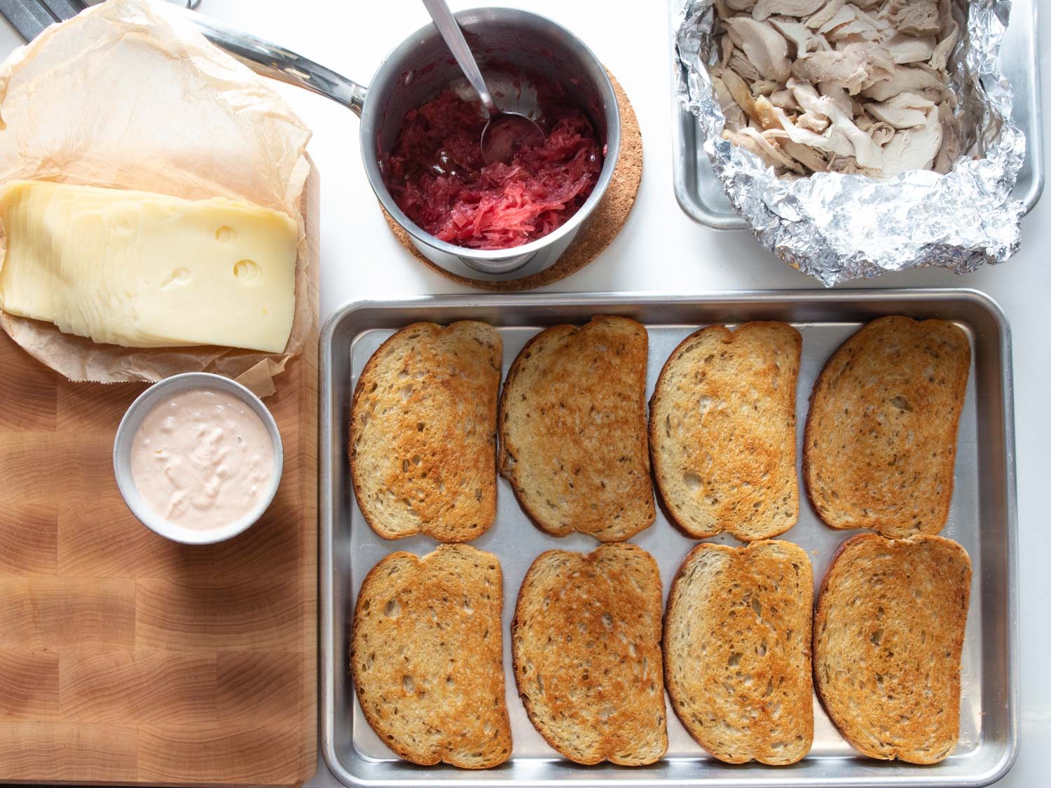 Overhead of components for turkey Reuben sandwiches: toasted rye bread, leftover roast turkey, cranberry-sauerkraut, Russian dressing, and Swiss cheese.