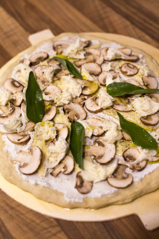 Close up photo of an uncooked white pizza topped with mushrooms and sage.