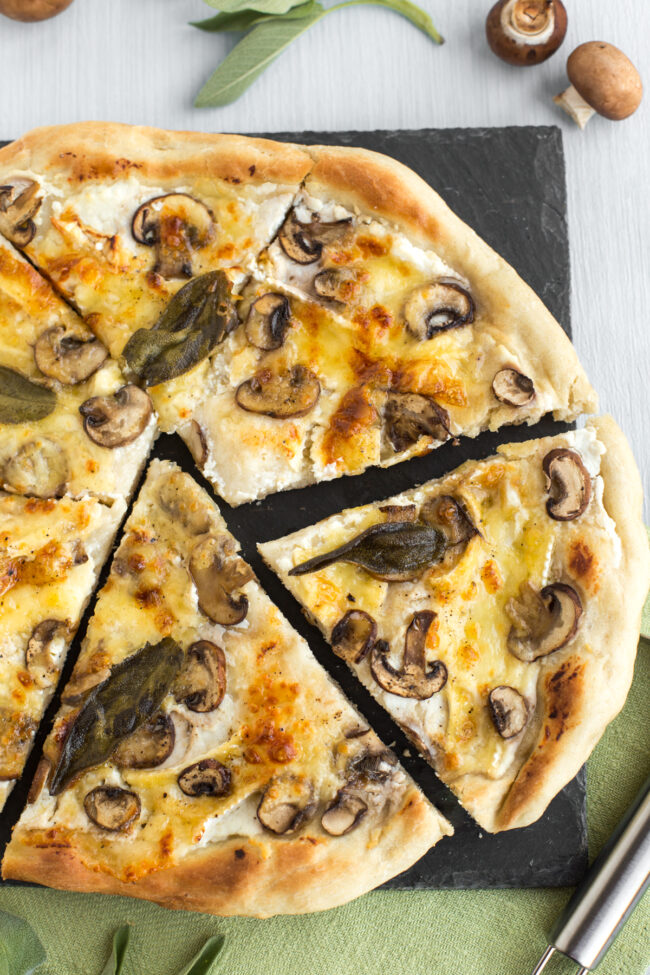 A vegetarian white pizza topped with mushrooms and sage leaves, cut into uneven slices.